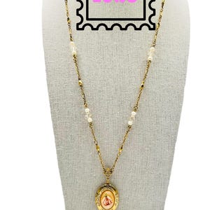 May include: Gold-coloured necklace with a pendant locket featuring a rose design. The necklace has a delicate chain with small pearls and accents. The image includes the text "1928" in a pink stamp-like frame.