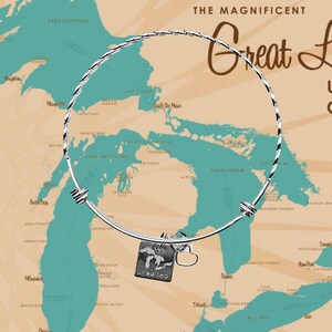 May include: A silver bangle bracelet with a charm depicting the Great Lakes and a heart. The bracelet is displayed against a map of the Great Lakes region.