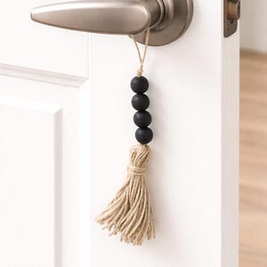 May include: A decorative door hanger featuring a tassel and black wooden beads. The tassel is made of natural-colored twine, and the beads are strung together with a neutral-colored cord. The door hanger is attached to a silver-colored door handle.