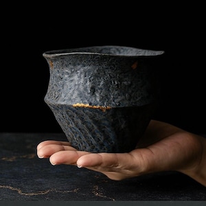 May include: A dark blue ceramic bowl with a textured surface and an irregular shape. The bowl has a gold band and is held in a hand. The bowl is a decorative item. The bowl's dimensions are in centimetres.