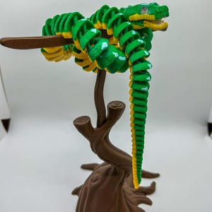 May include: A green and yellow articulated snake toy perched on a brown tree-shaped stand. The snake is coiled around a branch, with its head and tail hanging down. The toy is made of plastic and has a detailed design.