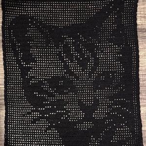 May include: A square, black crocheted blanket with a floral design. The blanket is made of a dark yarn, with a repeating pattern of open spaces that create the floral design. The blanket is likely handmade and has a textured appearance.
