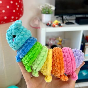 May include: A handmade, colorful crochet caterpillar toy. The caterpillar features a light blue head, a blue and green collar, and a body composed of segments in green, yellow, orange, pink, and purple. The toy is held in a hand.