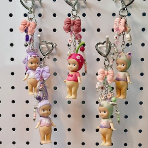 May include: Keychains featuring small figurines with various headwear, including a cherry, cat ears, and a purple bonnet. Each keychain has a pink bow and beaded accents. The figurines are displayed on a white pegboard, with boxes of Sonny Angel Mini Figures below.