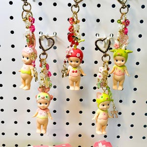 May include: Keychains featuring Sonny Angel mini figures from the Cherry Blossom Series. Each keychain has a gold heart-shaped clasp and decorative beads. The figures are wearing various fruit and flower-themed hats.