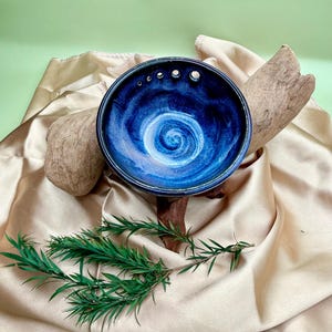 Puede incluir: Un cuenco de cerámica hecho a mano con un esmalte azul oscuro y un diseño en espiral blanco en el centro. El cuenco tiene varios agujeros pequeños cerca del borde. El cuenco se exhibe sobre una tela de satén dorada con madera flotante y ramitas verdes.