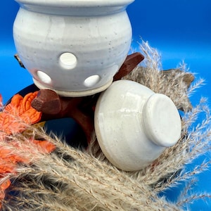 Pode incluir: Um queimador de óleo de cerâmica branca com uma tampa a condizer. O queimador tem uma forma redonda com vários orifícios e assenta numa base de madeira. O fundo inclui plantas secas e um fundo azul.