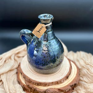 May include: A handcrafted, blue and black ceramic jug with a handle. The jug has a glossy, speckled finish and a small tag tied around the neck. It sits on a wooden slice, with a beige cloth in the background.