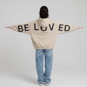 May include: A beige hoodie with the word "BELOVED" printed across the back in black block letters. The text "1 JOHN 3:1" is printed below the word "BELOVED". The person is wearing blue jeans.