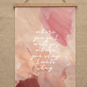 Puede incluir: Una impresión artística enmarcada con un diseño de acuarela en tonos rosa y melocotón. La impresión presenta la cita, "Where you go I will go, where you stay I will stay" en escritura blanca. El marco es de madera clara.