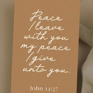 May include: A rectangular wall hanging with the text "Peace I leave with you, my peace I give unto you. John 14:27" in white script on a tan background. The top and bottom edges are framed with light wood.