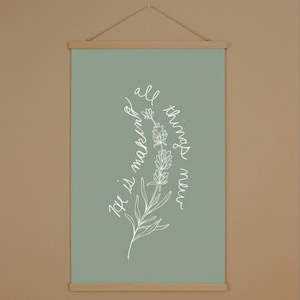 Puede incluir: Un cartel de pared verde salvia con una ilustración floral blanca y el texto manuscrito "He is making all things new". La obra de arte se sujeta con colgadores magnéticos de madera y una cuerda para colgar.