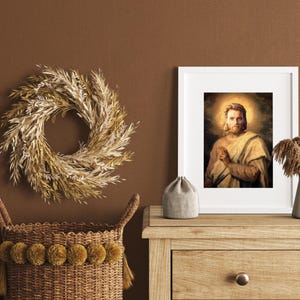 May include: A framed portrait of a man with a halo, a decorative wreath, a woven basket with pom-poms, a small gray pouch, and a white vase with dried plants. The items are arranged on a wooden table against a brown wall.
