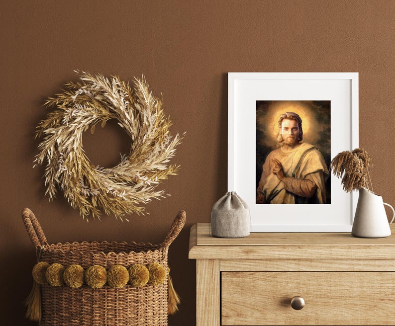 May include: A framed portrait of a man with a halo, a decorative wreath, a woven basket with pom-poms, a small gray pouch, and a white vase with dried plants. The items are arranged on a wooden table against a brown wall.