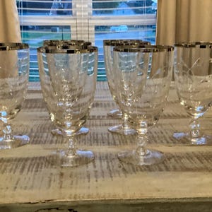 May include: Set of six clear glass goblets with silver rims and etched floral designs. The goblets have a classic shape with a stemmed base, perfect for serving water or wine at a formal dinner.