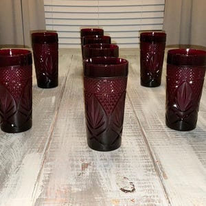 May include: A set of six deep red glass tumblers featuring a textured geometric pattern on the upper half and a pressed leaf-like design on the lower half. Each cylindrical drinking glass has a smooth rim and a consistent dark ruby color throughout.