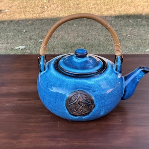 May include: A blue ceramic teapot with a wooden handle and a dark brown emblem. The teapot has a textured surface and a spout. The handle is made of woven rattan. The teapot is on a wooden surface.