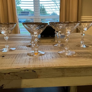 May include: Six clear champagne glasses with gold rims are arranged on a distressed wooden table. The glasses have ornate stems and a classic coupe shape. The table is light-colored with visible wood grain.