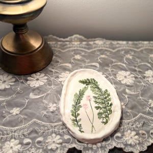 Könnte beinhalten: Eine kleine, cremefarbene Keramikschale mit handgemaltem botanischem Design. Das Design zeigt grüne Farne und eine rosa Blume. Die Schale steht auf einer weißen Spitzentischdecke mit floralen Stickereien.
