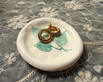 Puede incluir: Un pequeño plato de cerámica blanca con un diseño de hojas en verde azulado y pendientes de aro dorados. El plato tiene una forma orgánica y está sobre una superficie de encaje gris.