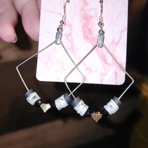 May include: Silver-coloured, geometric earrings with a square design. Each earring features a silver hook, a square frame, and a series of black and white beads, with a small gold heart charm in the centre. The earrings are displayed on a pink card.