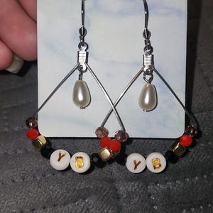 May include: Silver earrings with a teardrop pearl and a beaded hoop design. The hoop features red, black, gold, and clear beads, with white letter beads spelling "YB".