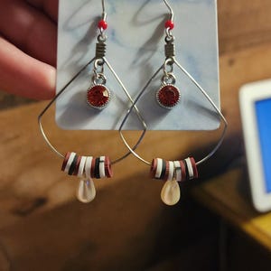 May include: A pair of handmade earrings featuring silver-tone hoops with a geometric design. Each earring has a red gemstone, red beads, and a teardrop-shaped charm. The earrings are displayed on a light blue card.