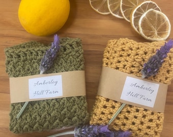 May include: An overhead shot features two crocheted washcloths, one olive green and one mustard yellow, each wrapped with a brown paper band that reads "Amberley Hill Farm." The cloths are adorned with lavender sprigs, alongside lemons and dried lemon slices.