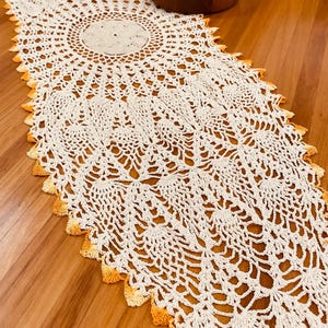 May include: Cream-coloured, crocheted table runner with a detailed openwork design. The runner has a central circular motif and a scalloped edge with a gradient of yellow-orange. The runner is displayed on a wooden surface.