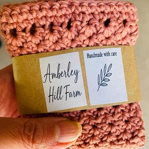 May include: A folded, coral-colored, crocheted item with a brown paper band. The band features the text "Amberley Hill Farm" and "Handmade with care" with a leaf illustration. The item is held in a hand.