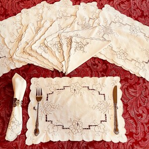 May include: A set of off-white rectangular placemats and matching napkins featuring intricate floral embroidery and scalloped edges. One placemat is laid out with a silver fork and knife, while a napkin is held in a metallic napkin ring.