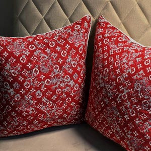 Puede incluir: Dos cojines decorativos rojos con borde blanco. Los cojines presentan un patrón repetitivo de diseños florales y de logotipos blancos. Los cojines descansan sobre una superficie beige acolchada.