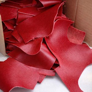 May include: A cardboard box overflowing with irregularly shaped pieces of red leather. The leather has a textured surface and varying sizes. The pieces are scattered, with some spilling out of the box.