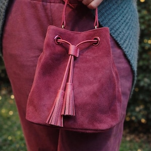 May include: A burgundy suede bucket bag with a drawstring closure and matching tassels. The bag has a structured shape and a small handle. The bag is held by a person wearing burgundy pants and a gray sweater.