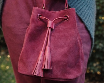 May include: A burgundy suede bucket bag with a drawstring closure and matching tassels. The bag has a structured shape and a small handle. The bag is held by a person wearing burgundy pants and a gray sweater.