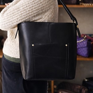 May include: A black leather tote bag with a rectangular shape and a top handle. The bag has a front pocket with a gold-colored stud. Other bags are visible on shelves in the background.
