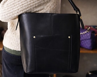 May include: A black leather tote bag with a rectangular shape and a top handle. The bag has a front pocket with a gold-colored stud. Other bags are visible on shelves in the background.
