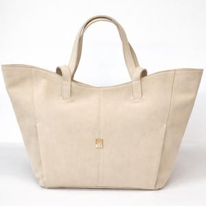 May include: A beige tote bag with two top handles and a wide, open-top design. The bag features a central vertical seam and a small, square gold-toned metal emblem centered on the front panel. The material has a smooth, matte, uniform finish.