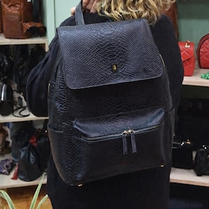 Op de afbeelding: Zwarte rugzak met een textuur die op slangenleer lijkt. De tas heeft een klepsluiting, een voorvak met ritssluiting en goudkleurige ritsen. De rugzak wordt gedragen.