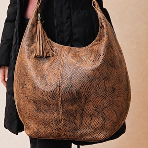 May include: A large brown hobo bag with a snakeskin pattern. The bag has a curved shape, a long shoulder strap, and a decorative tassel. The bag is held by a person wearing a black coat.