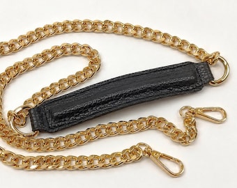 May include: Gold-tone metal chain strap with a black leather shoulder pad. The chain has a classic curb link design and includes two gold-tone lobster clasps for easy attachment to a bag.