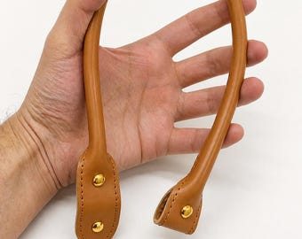 May include: A tan leather handbag handle with gold-colored rivets. The handle is curved and appears to be new. The handle is held in a person's hand against a white background.