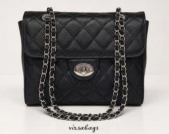 May include: A black quilted handbag with a textured finish, featuring a silver-tone metal turn-lock closure and a dual chain strap interwoven with black material. The text virsabags is printed in black below the bag.