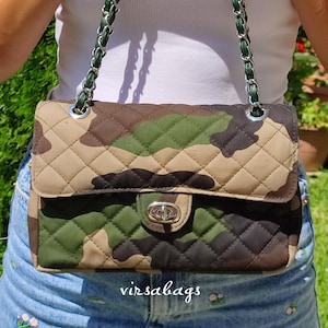 Peut inclure: Sac à bandoulière matelassé à motif camouflage avec une bandoulière chaîne argentée. Le sac est doté d'une fermeture à rabat avec un fermoir argenté. Le sac est dans les tons de vert, de marron et de beige. Le texte "virsabags" est visible.