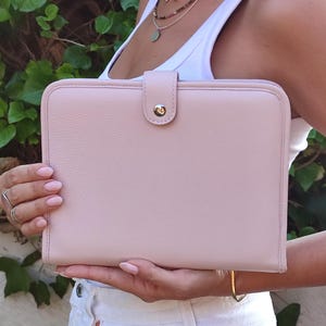 Puede incluir: Un bolso de mano rectangular de color rosa pálido con cierre de botón a presión. El bolso se sostiene frente a una persona que lleva una camiseta sin mangas blanca y vaqueros blancos. El fondo es follaje verde.