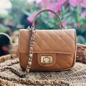 Puede incluir: Un bolso acolchado de color tostado con un patrón de costuras en forma de diamante, un asa superior y un cierre giratorio de metal plateado. El bolso incluye una correa larga para el hombro hecha de una cadena plateada entrelazada con material tostado.