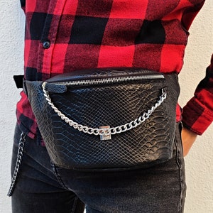 May include: Black fanny pack with a snakeskin texture and silver chain detail. The bag has a zipper closure and a silver clasp. The fanny pack is worn around the waist.