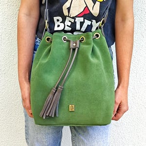 May include: A green suede bucket bag with a drawstring closure, brown leather straps, and gold-tone metal hardware. The bag features a small rectangular gold logo plaque on the front and two decorative leather tassels hanging from the drawstring.