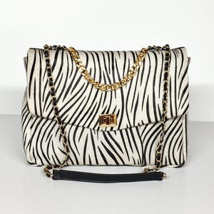 May include: A rectangular shoulder bag featuring a black and white zebra stripe pattern. The bag includes a gold-tone metal turn-lock closure on the front flap and a shoulder strap composed of gold-tone metal chains interwoven with black material.