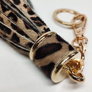Peut inclure: Un porte-clés composé d'un long pompon à plusieurs brins avec un motif imprimé léopard marron et noir. Le pompon est fixé par un capuchon en métal doré et est attaché à un anneau fendu en métal doré et un fermoir mousqueton.